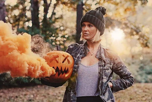 Woman in beanie carrying pumpkin spilling orange smoke in autumn woods